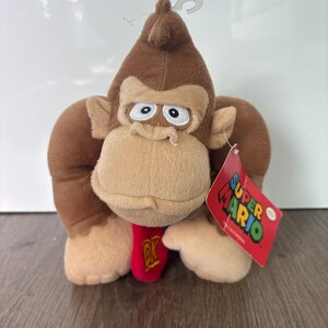 Könnte beinhalten: Ein braunes Gorilla-Plüschtier mit beigem Gesicht, weißen Augen und einer roten Krawatte mit den gelben Buchstaben DK. Das Spielzeug hat eine weiche Textur und ein rotes Pappschild an der Seite mit der Aufschrift Super Mario in bunten Buchstaben.
