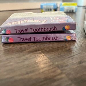 Puede incluir: Dos paquetes de cepillo de dientes de viaje de color morado y blanco con el texto "Travel Toothbrush" y "Popsies Travel Toothbrush" en la parte delantera.