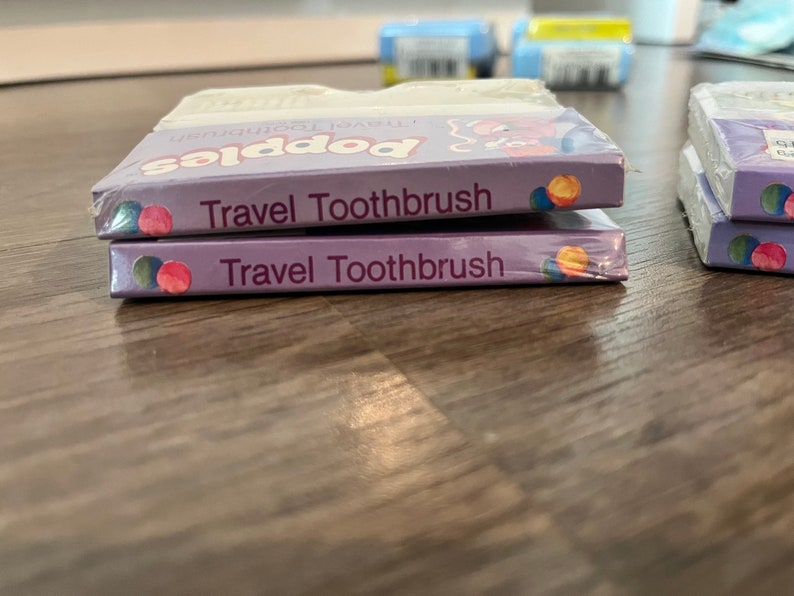 Puede incluir: Dos paquetes de cepillo de dientes de viaje de color morado y blanco con el texto "Travel Toothbrush" y "Popsies Travel Toothbrush" en la parte delantera.