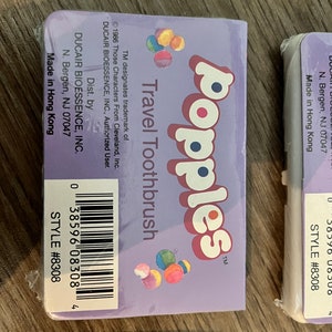 Puede incluir: Un paquete de cepillo de dientes de viaje morado con el texto "Popples Travel Toothbrush" y una imagen colorida de los personajes de Popples. El paquete tambi&eacute;n incluye el texto "Dist. by DUCAIR BIOESSENCE, INC. N. Bergen, NJ 07047 Made in Hong Kong" y un c&oacute;digo de barras.