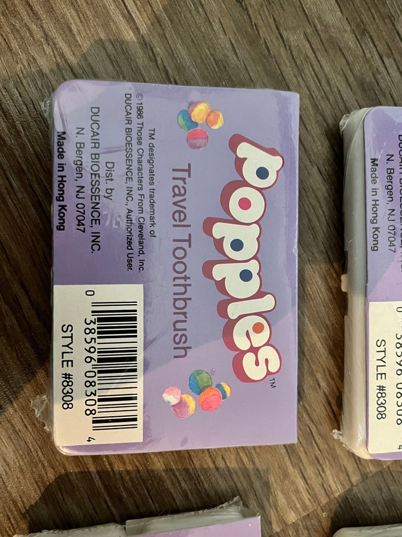 Puede incluir: Un paquete de cepillo de dientes de viaje morado con el texto "Popples Travel Toothbrush" y una imagen colorida de los personajes de Popples. El paquete tambi&eacute;n incluye el texto "Dist. by DUCAIR BIOESSENCE, INC. N. Bergen, NJ 07047 Made in Hong Kong" y un c&oacute;digo de barras.