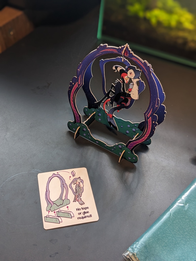 May include: A 3D papercraft model of two figures standing in a purple and blue archway. The figures are a person with red hair and a person with blue hair. The model is made of cardboard and is assembled without glue or tape. The instructions for assembling the model are shown on a separate card.