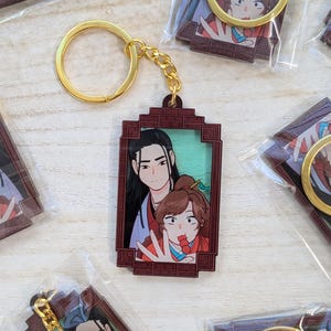 May include: A brown and gold keychain with a picture of two people in a frame. The picture shows a person with long black hair and a person with brown hair. The person with brown hair is wearing a red and gold outfit and is holding a red lollipop. The keychain has a gold ring and a chain.