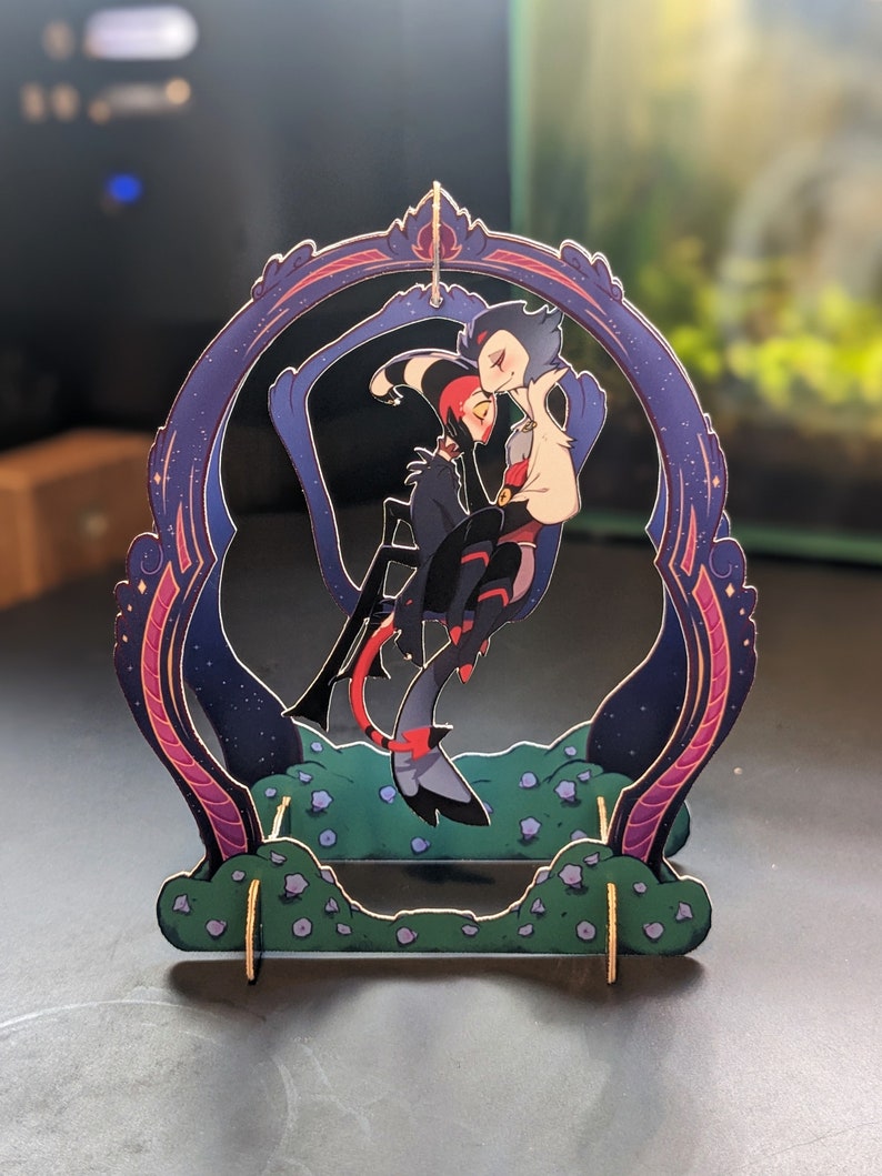May include: A wooden stand with a decorative frame featuring two cartoon characters in a romantic embrace. The characters are a red and black demon with horns and a blue and white angel with wings. The stand has a starry night sky background and a green base with small flowers.