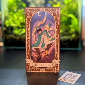 May include: A tarot card with a brown frame and ornate details. The card depicts two figures in blue and green clothing, holding scrolls, against a purple and pink background. The text "THE FUTURE" is printed on the card.