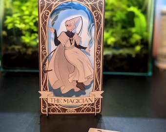 May include: A tarot card with a gold frame and a blue and white illustration of a magician. The card reads "The Magician" in a banner at the bottom.
