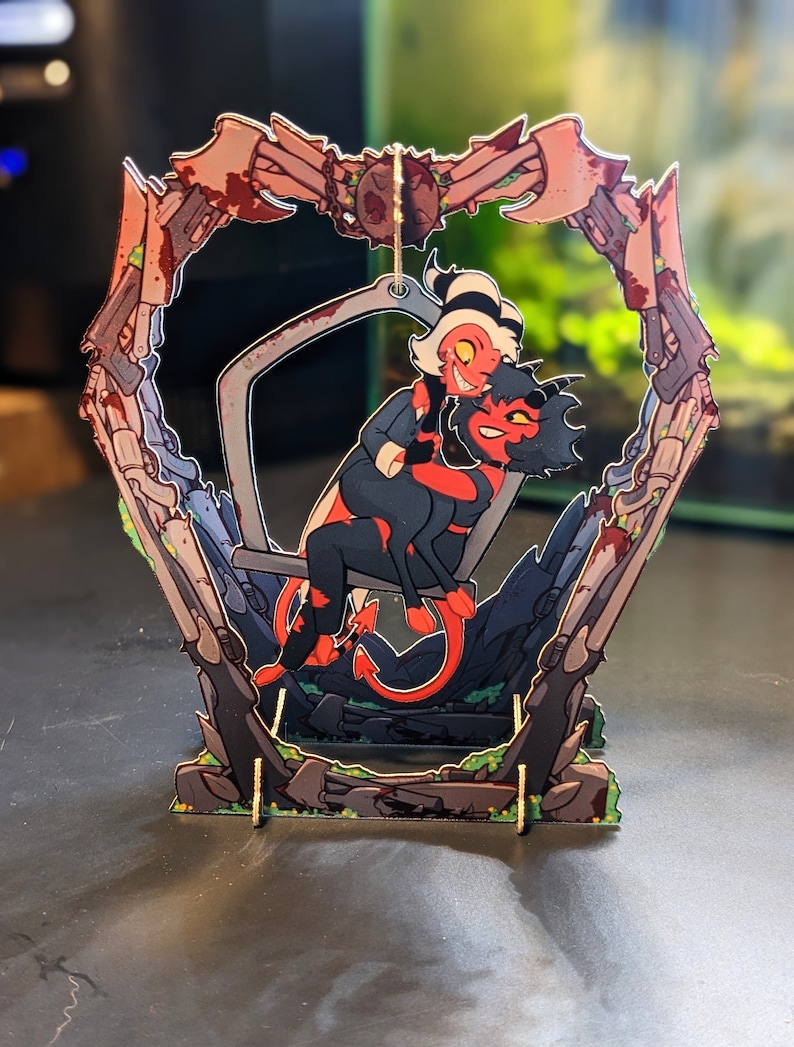 May include: A red and black cartoon character with horns and a tail is sitting on a swing with another cartoon character. The swing is suspended from a metal frame with a heart-shaped opening. The frame is decorated with a bloody design.