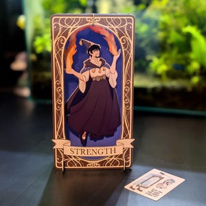 Puede incluir: Una carta del tarot con fondo azul y ribete dorado. La carta representa a una persona con una túnica oscura que sostiene llamas en cada mano. La carta está etiquetada como "FUERZA".