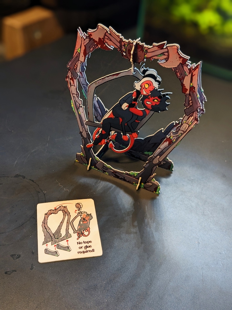May include: A 3D papercraft model of two cartoon characters, a red demon and a white demon, sitting on a swing. The swing is made of brown cardboard and is shaped like a heart. The model is assembled and ready to display. The instructions on the card say "No tape or glue required!"