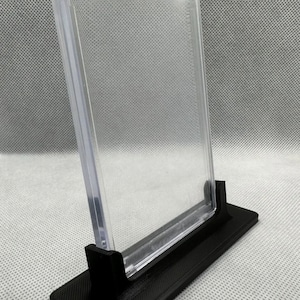 May include: A clear plastic display case with a black base. The case is designed to hold and display a collectible card or other small item.