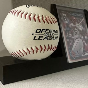May include: A white baseball with red stitching and the text "OFFICIAL LEAGUE OLB3" is displayed on a black stand. A baseball card with a player in a red uniform is displayed in a black frame to the right of the baseball.