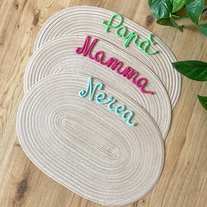 Può includere: Tre tovagliette ovali in tessuto beige impilate verticalmente. Ogni tovaglietta presenta un nome in corsivo realizzato con filo in rilievo: quella in alto in verde brillante riporta Papa, quella centrale in rosa acceso Mamma e quella in basso in azzurro Nerea.