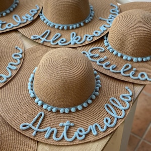 Può includere: Diversi cappelli di paglia marrone con finiture azzurre e nomi personalizzati in corsivo. I cappelli hanno una tesa larga e una corona rotonda.