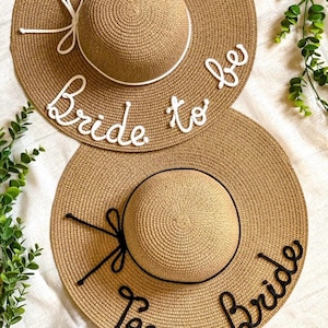 Può includere: Due cappelli di paglia con tesa larga. Uno ha le parole "Bride to be" in corsivo bianco, l'altro le parole "Team Bride" in corsivo nero. Entrambi i cappelli hanno una corona rotonda e un fiocco decorativo.