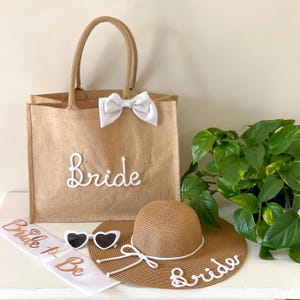 Op de afbeelding: Een bruids cadeau set met een bruine jute tote bag met "Bride" in witte letters, een witte strik, een strohoed met brede rand met "Bride" in witte letters, hartvormige zonnebrillen en een witte sjerp met "Bride to Be".