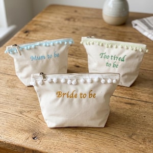 Può includere: Tre pochette in tela color bianco sporco con cerniera e bordo a pompon lungo il bordo superiore. La parte anteriore di ogni pochette presenta un testo ricamato: azzurro Mum to be, verde Too tired to be e oro Bride to be, con chiusura a zip.