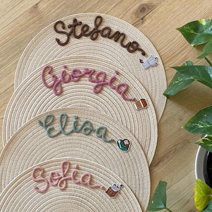 Op de afbeelding: Vier geweven placemats met geborduurde namen: Stefano, Giorgia, Elisa en Sofia. Elke naam heeft een andere kleur: bruin, roze, teal en roze. Kleine decoratieve spelden zijn aan elke placemat bevestigd. Een groene plant is zichtbaar op de achtergrond.
