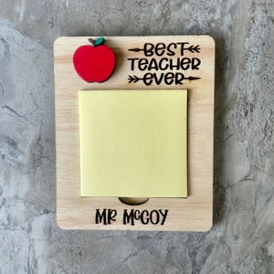May include: Wooden notepad with a red apple and the text "Best Teacher Ever" with an arrow pointing to the right. The notepad has a yellow sticky note and the name "Mr. McCoy" is written below the sticky note.