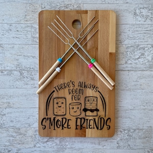 Puede incluir: Una tabla de cortar de madera con temática de s'mores. La tabla tiene un diseño negro que dice "Siempre hay espacio para más amigos". El diseño incluye un malvavisco, una barra de chocolate y una galleta graham. La tabla también tiene cuatro pinchos de metal con mangos de madera.
