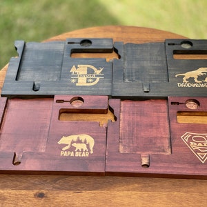 May include: Four wooden phone stands with different designs. The first stand has a black stain and is engraved with a tree, a deer, and the name "David". The second stand is also black and has a dinosaur and the text "Daddy-saurus". The third stand is a reddish-brown color and has a bear and the text "Papa Bear". The fourth stand is also reddish-brown and has a Superman logo with the text "Dad".