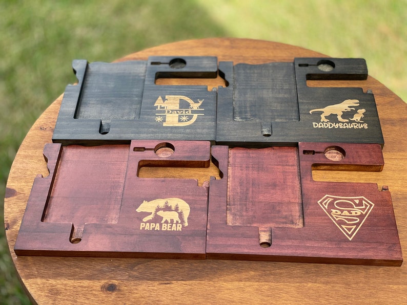 May include: Four wooden phone stands with different designs. The first stand has a black stain and is engraved with a tree, a deer, and the name "David". The second stand is also black and has a dinosaur and the text "Daddy-saurus". The third stand is a reddish-brown color and has a bear and the text "Papa Bear". The fourth stand is also reddish-brown and has a Superman logo with the text "Dad".