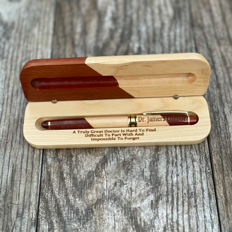 May include: A wooden pen box with a red and natural wood finish. The box is open and a gold pen with a red and natural wood barrel is inside. The box is engraved with the text "Dr. Jamers como A Truly Great Doctor Is Hard To Find Difficult To Part With And Impossible To Forget".