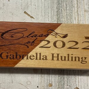 May include: A wooden plaque with a brown and light wood finish. The plaque is engraved with the text "Class of 2022" and "Gabriella Huling".