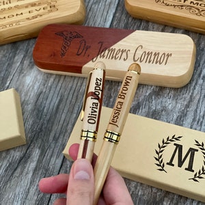 May include: Two wooden pens with gold accents and personalized names, "Olivia Lopez" and "Jessica Brown", are held in a hand. The pens are in front of a wooden box with the engraved text "Dr. Jamers Connor".