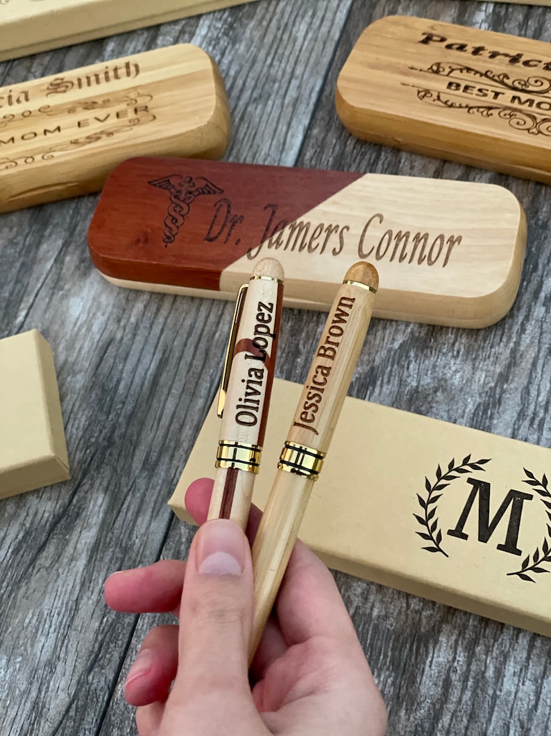 May include: Two wooden pens with gold accents and personalized names, "Olivia Lopez" and "Jessica Brown", are held in a hand. The pens are in front of a wooden box with the engraved text "Dr. Jamers Connor".