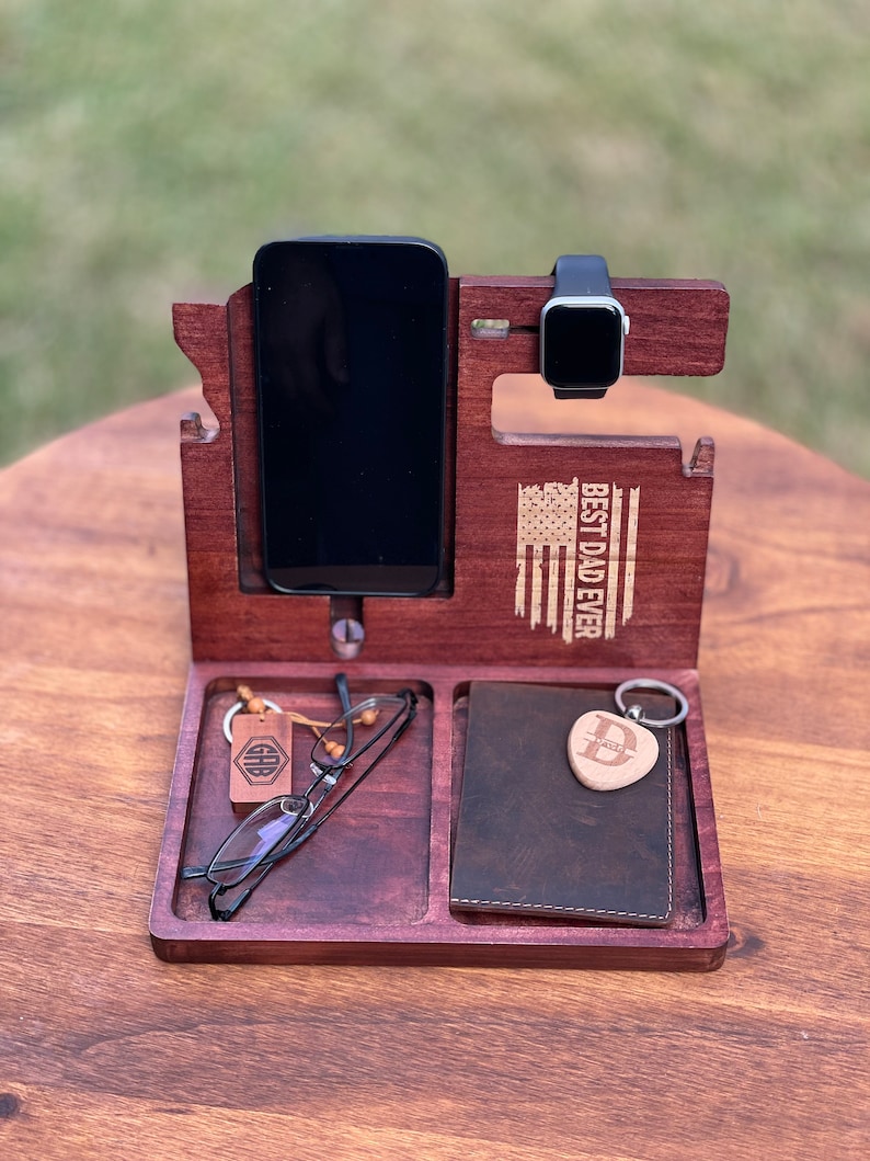 May include: A wooden phone stand with a phone, watch, glasses, wallet, and keys. The stand is engraved with the text "Best Dad Ever" and a small American flag.