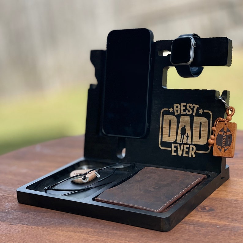 May include: Black wooden phone stand with a phone, watch, glasses, and wallet slots. It has the words "Best Dad Ever" engraved on the front.