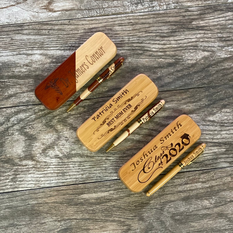 May include: Three wooden boxes with engraved personalized messages and pens. The boxes are made of light brown wood with a natural wood grain. The pens are gold and black with a wood finish. The messages on the boxes are "Jamers Connor", "Patricia Smith BEST MOM EVER", and "Joshua Smith Class of 2020".