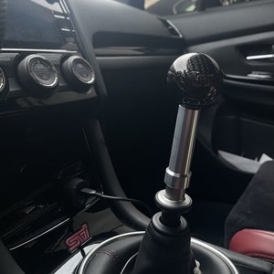 May include: A close-up of a black carbon fiber gear shift knob with a silver extension. The knob is installed in a car with a black leather interior and a red "STI" logo.