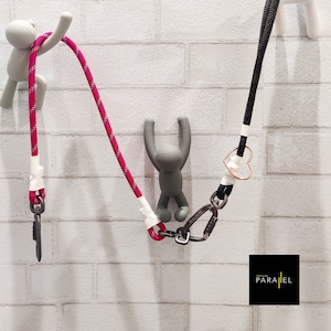 May include: Three gray and white plastic hooks shaped like people, hanging on a white brick wall. Each hook has a different colored rope attached to it: pink, black, and white. The ropes are attached to a carabiner clip.