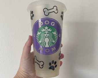 May include: A clear plastic tumbler with a green straw, featuring a purple and green logo with the words "DOG MOM". The tumbler is decorated with black paw prints and bone graphics.