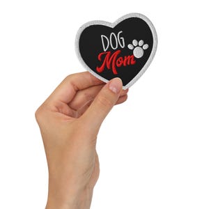 May include: Heart-shaped patch with a black background and white stitching. The patch features the words "DOG Mom" in white and red, along with a white paw print. The patch is held in a hand.