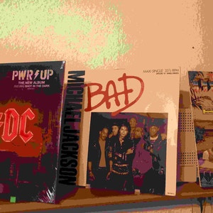 Puede incluir: Tres discos de vinilo en un estante. El primer disco es el álbum "PWR UP" de AC/DC, el segundo es el álbum "BAD" de Michael Jackson, y el tercero es un disco con el texto "TWILIGHT ZONE" en la portada.