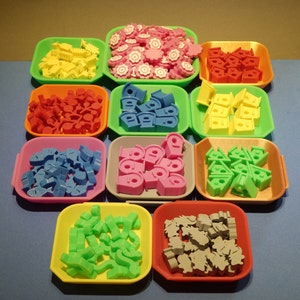 Könnte beinhalten: Sortiment aus bunten Plastik-Spielsteinen in verschiedenen Formen und Größen. Die Spielsteine sind in zehn kleinen Behältern sortiert, jeder in einer anderen Farbe.