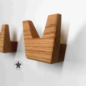 May include: A set of wooden wall hooks, each featuring a distinctive V-shaped design. The hooks are crafted from light brown wood and are mounted on a white wall. The hooks are part of a set, with a turtle logo.