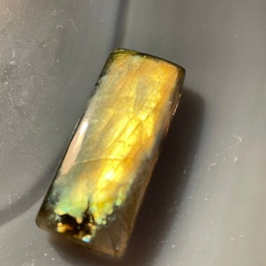 Puede incluir: Una piedra preciosa de labradorita rectangular e iridiscente con un tono dorado y destellos de verde y azul.