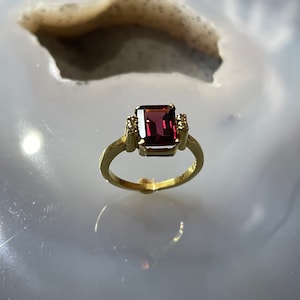 Op de afbeelding: Een gouden ring met een grote, donkerrode, smaragd geslepen edelsteen, omringd door kleinere, heldere edelstenen.