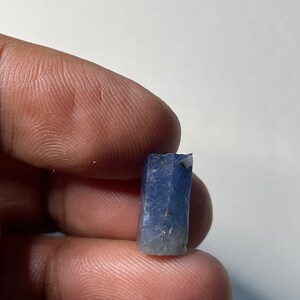 Puede incluir: Un pequeño cristal rectangular de color azul con una superficie rugosa y texturizada. El cristal se sostiene entre dos dedos sobre un fondo blanco. El color del cristal varía de azul claro a oscuro.
