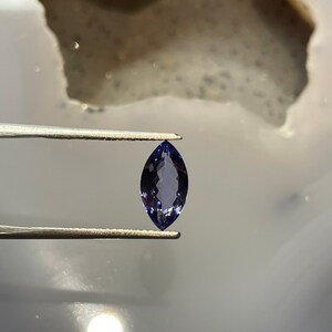 May include: A marquise-shaped gemstone with a deep purple hue. The gemstone is held by tweezers against a white background.