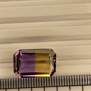 Puede incluir: Una piedra preciosa rectangular de color púrpura y amarillo, que mide aproximadamente 1,5 cm de largo, se muestra en una regla.