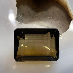 Puede incluir: Una piedra preciosa de cuarzo ahumado con un corte rectangular y un color marrón oscuro. La piedra preciosa está colocada sobre un fondo blanco.