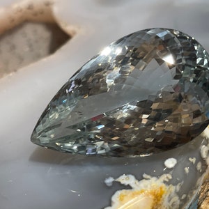 May include: A pear-shaped, light green gemstone with a faceted cut. The gemstone is resting on a white and brown surface.