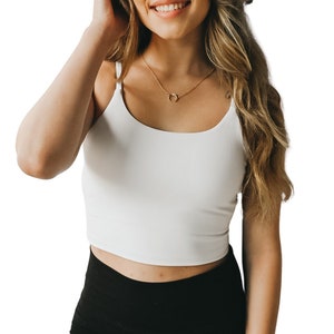 May include: A woman with long, wavy blonde hair is wearing a white tank top and black leggings. She is smiling and has a gold necklace with a crescent moon pendant.