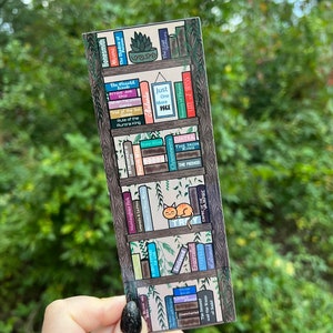 May include: A bookmark featuring a bookshelf with a variety of colorful books, including titles like "The Wegulil", "Rule of the Queen", "Just One More Page", "The Iron King", and "The Method". A small orange cat sits on a shelf, looking at the viewer.