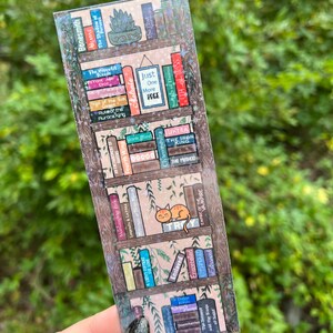以下が含まれることがあります： 様々な本のタイトルが書かれた本棚が描かれたホログラフィックブックマーク。タイトルには、「The Iron King」、「Stone Blind」、「Just One More Page」、「The Shadow of Perseus」などがあります。緑色の植物と一緒に、小さなオレンジ色の猫が棚の上に座っています。
