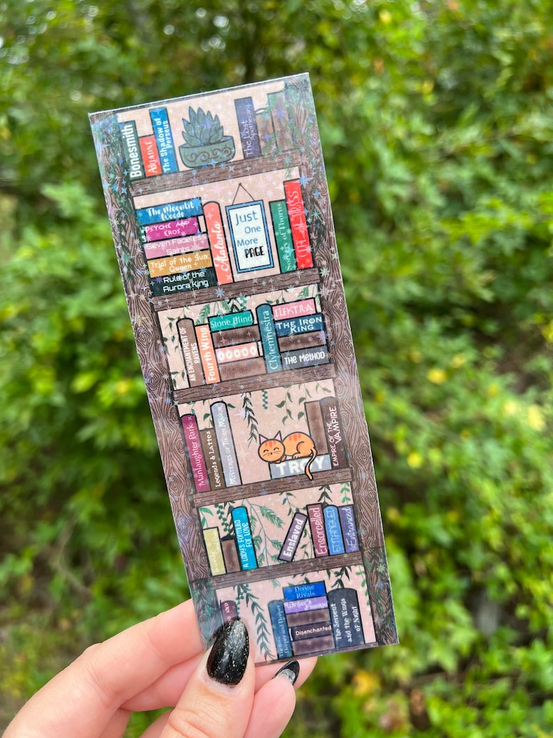 以下が含まれることがあります： 様々な本のタイトルが書かれた本棚が描かれたホログラフィックブックマーク。タイトルには、「The Iron King」、「Stone Blind」、「Just One More Page」、「The Shadow of Perseus」などがあります。緑色の植物と一緒に、小さなオレンジ色の猫が棚の上に座っています。
